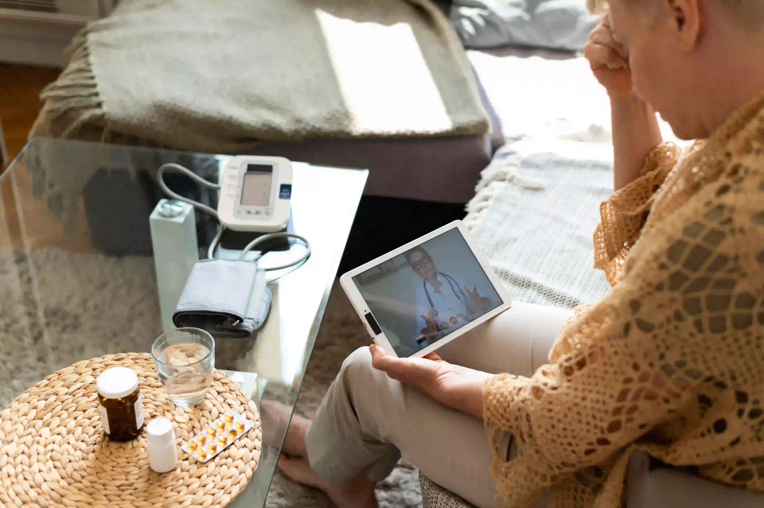Older woman having an online medical consultation on a tablet, with medicine and blood pressure monitor on table.