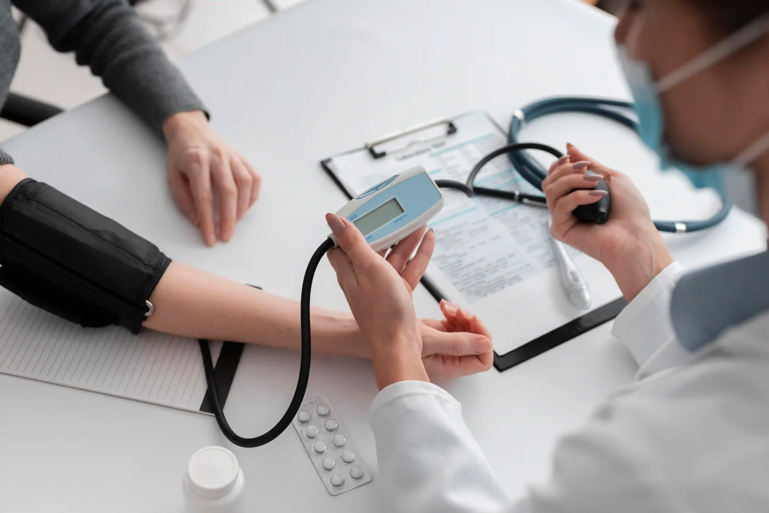 Doctor measuring patient’s blood pressure and reviewing medical documents at a clinic table.