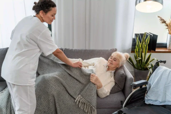 A healthcare worker in a white uniform covers an elderly woman with a blanket, who is lying on a sofa in a living room.