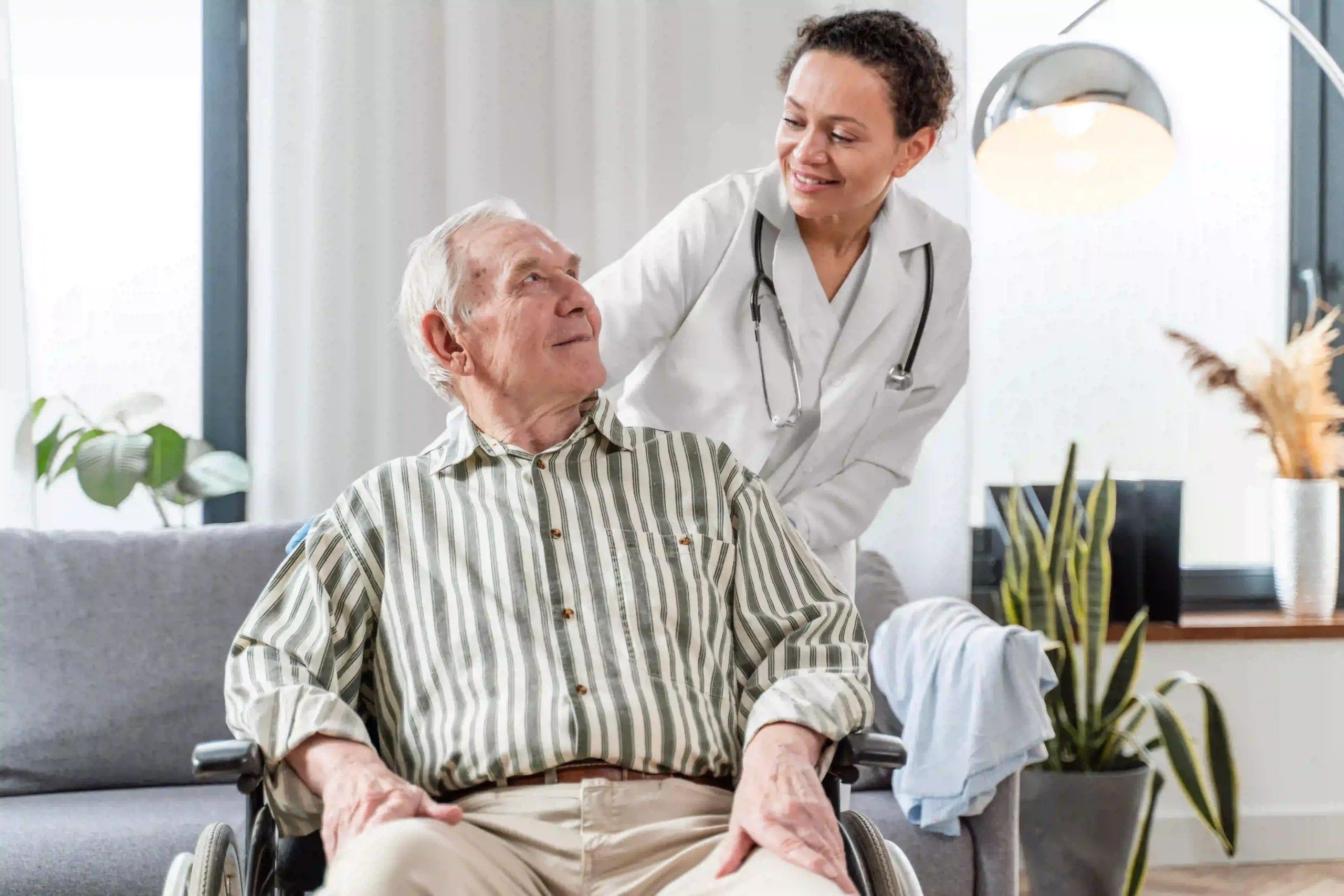 Female doctor talking warmly to an older man in a wheelchair during a home medical visit.