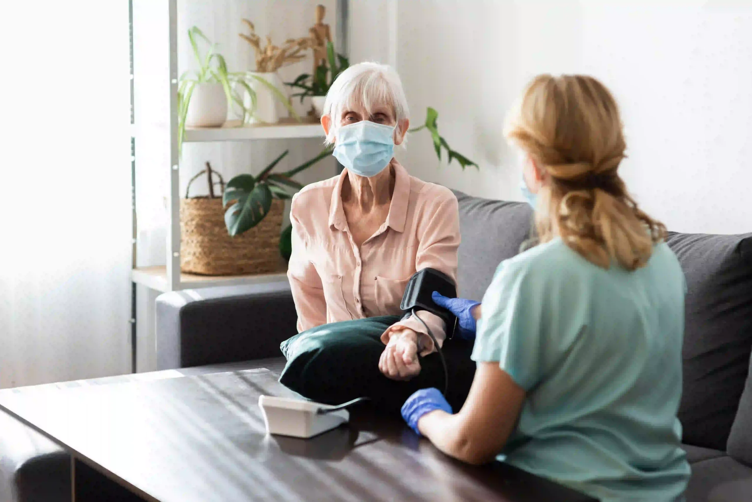 An elderly woman wearing a medical mask sits on a sofa as a healthcare worker in a blue uniform and gloves checks her blood pressure.
