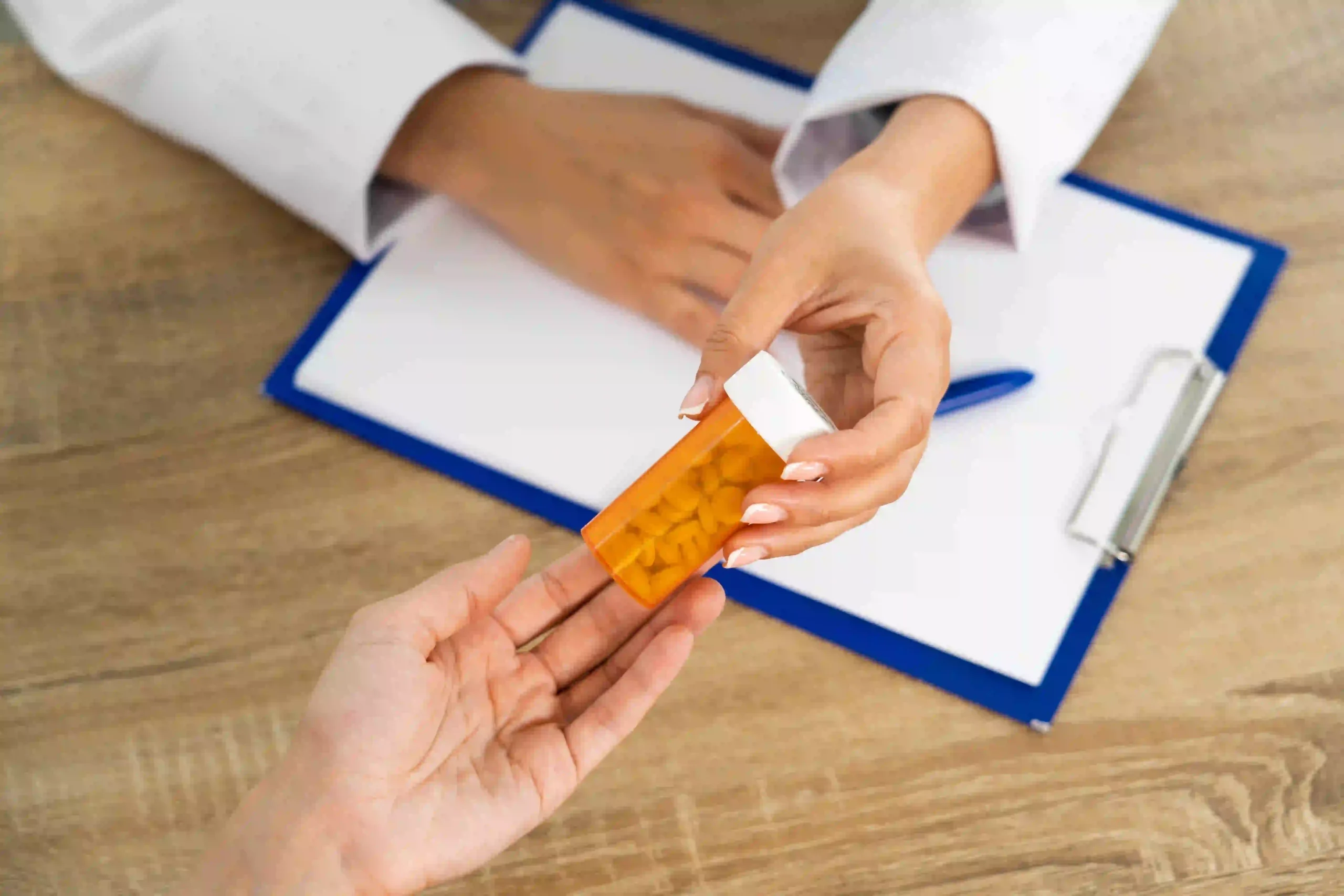 Doctor handing a prescription pill bottle to a patient, clipboard and pen on the wooden table.