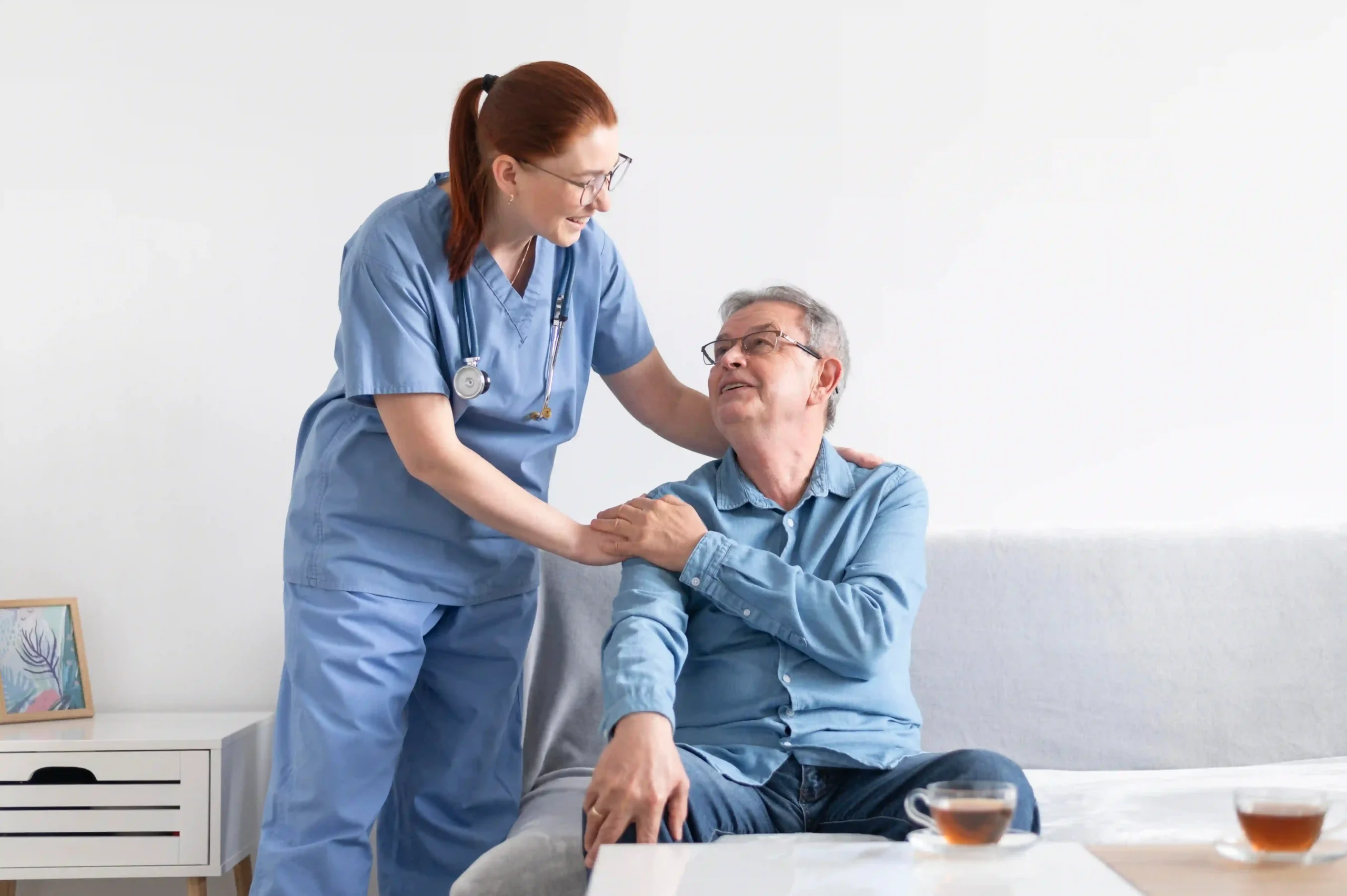 Smiling nurse supporting and reassuring an elderly man sitting on a couch during a home visit.