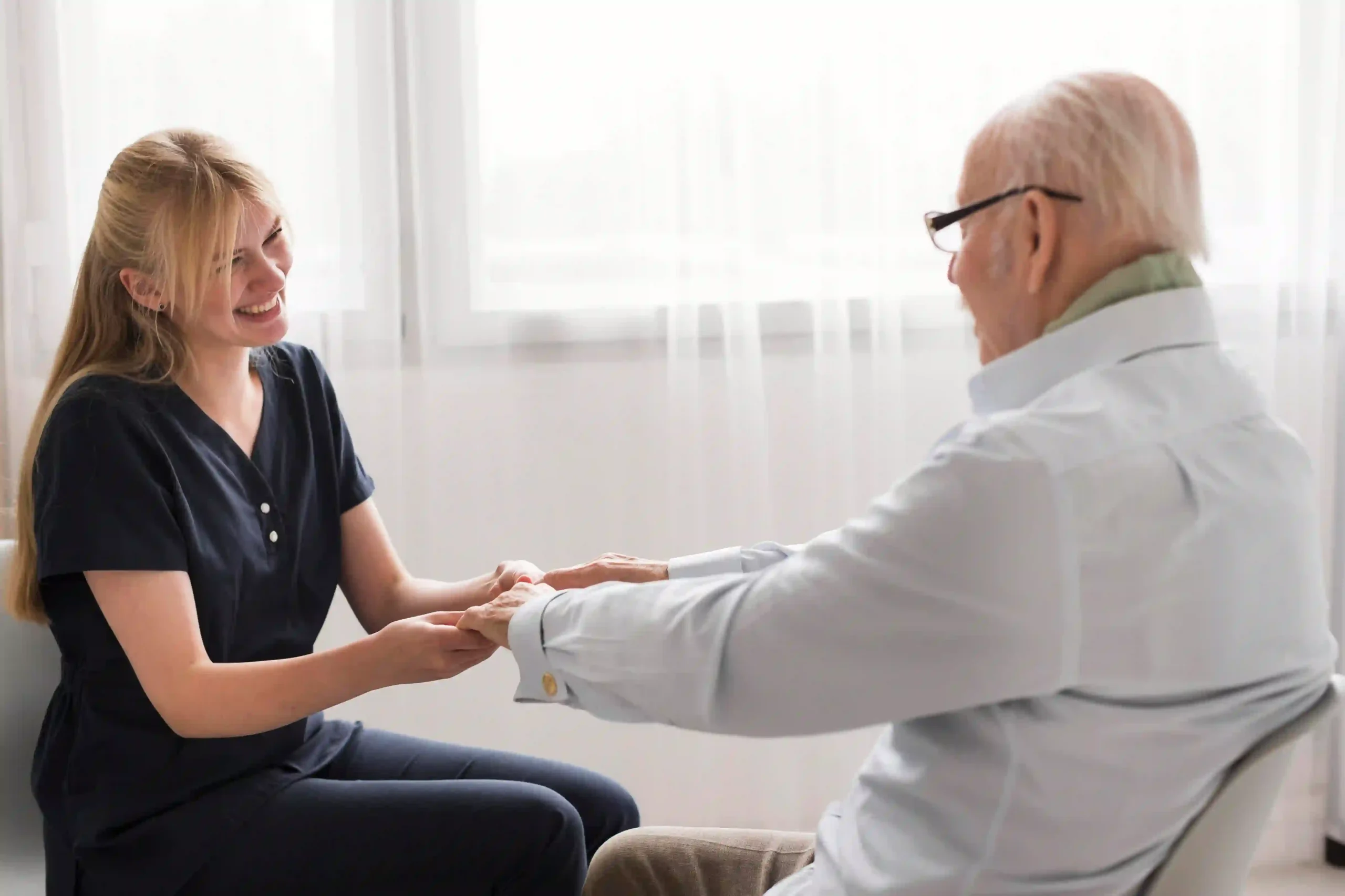 A smiling caregiver holds hands with an elderly man in a bright assisted living room.