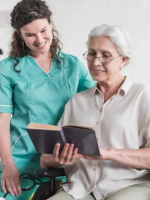 Smiling nurse assisting a senior woman in a wheelchair with reading a book in a comforting care environment.