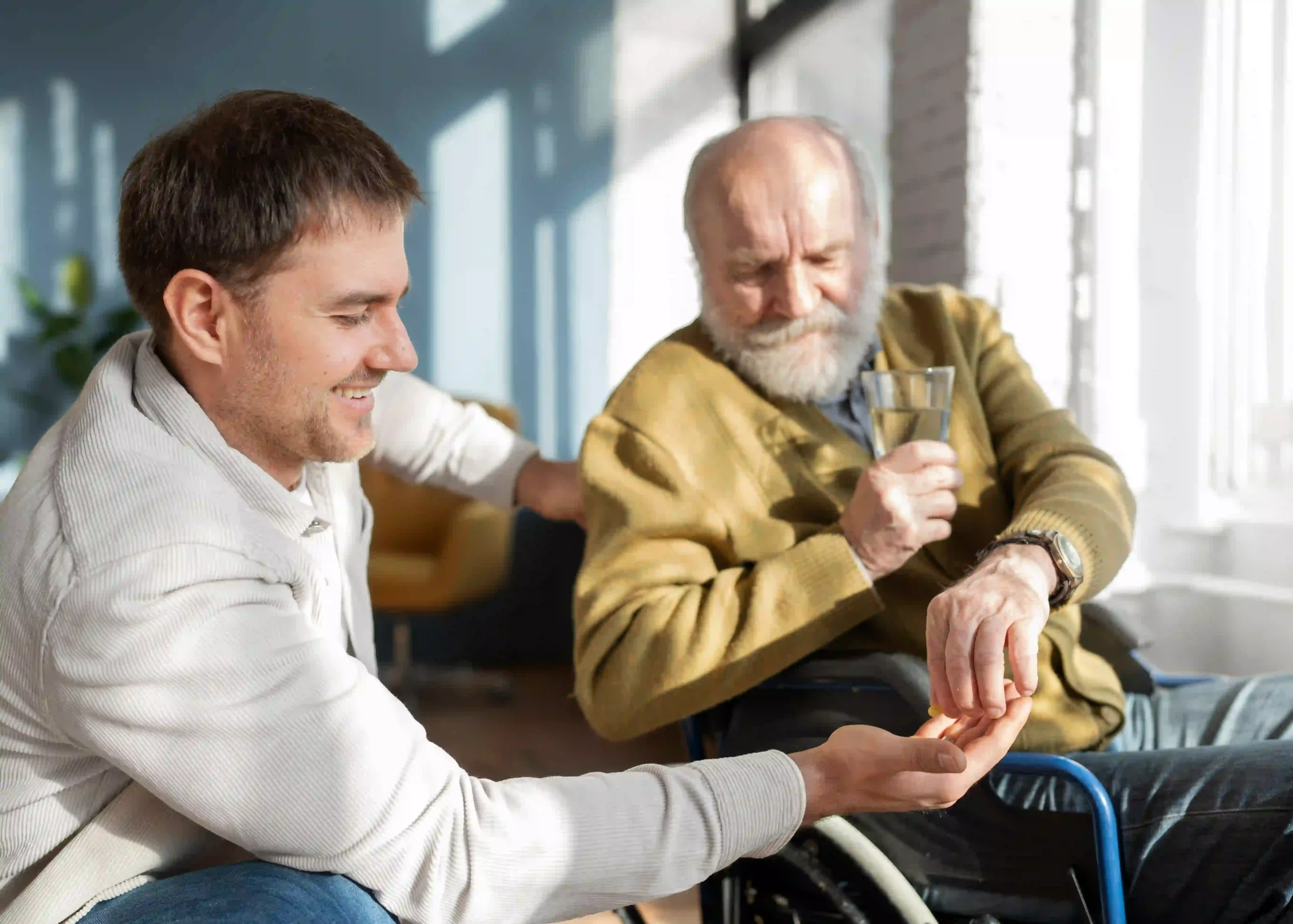 Elderly man in a wheelchair receiving assistance with medication from a younger man at home.