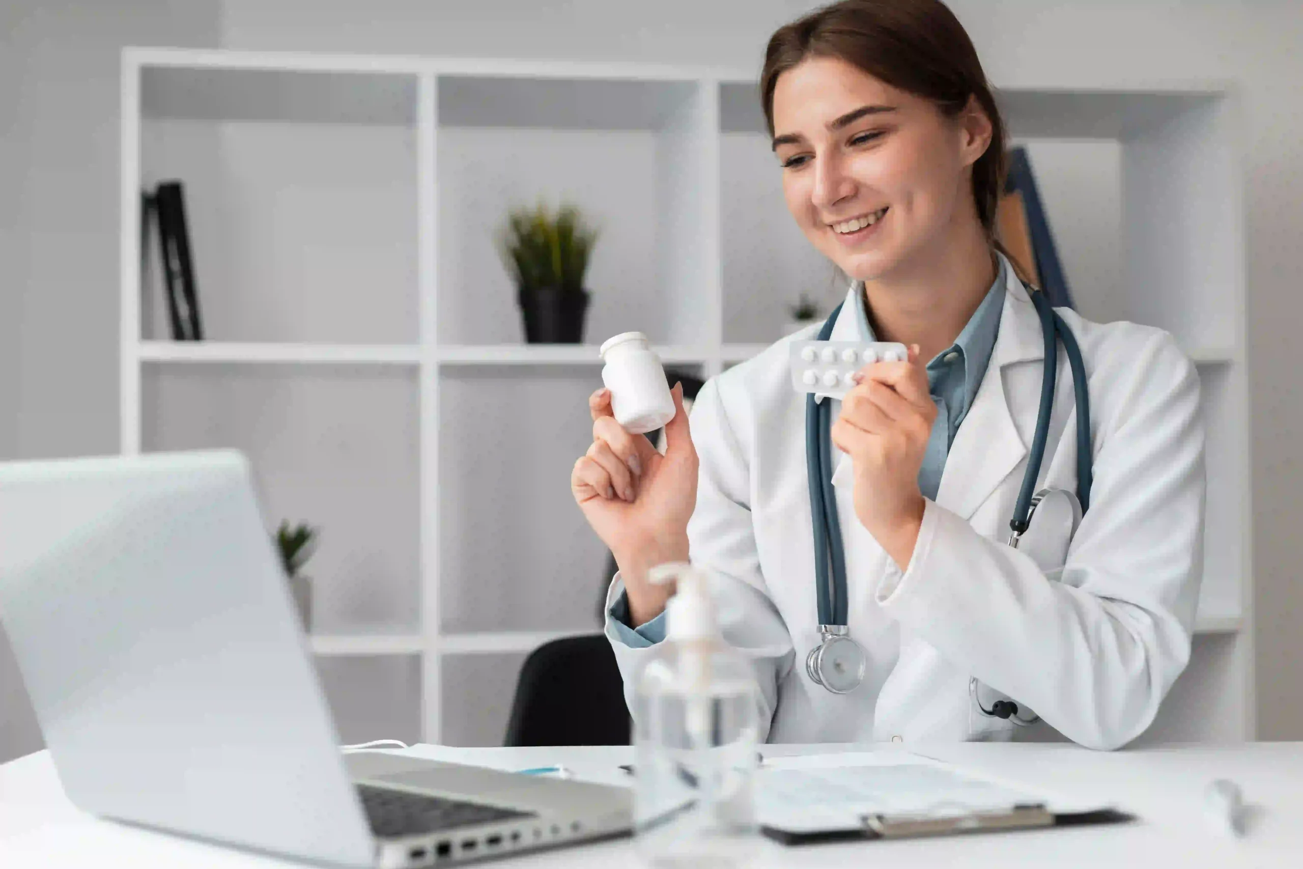 A young female medic in a white lab coat and a stethoscope smiles, holding a bottle of pills and a blister pack, while sitting in front of a laptop.