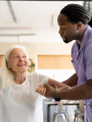 Male caregiver helping an older woman stand up safely at home with a supportive smile.