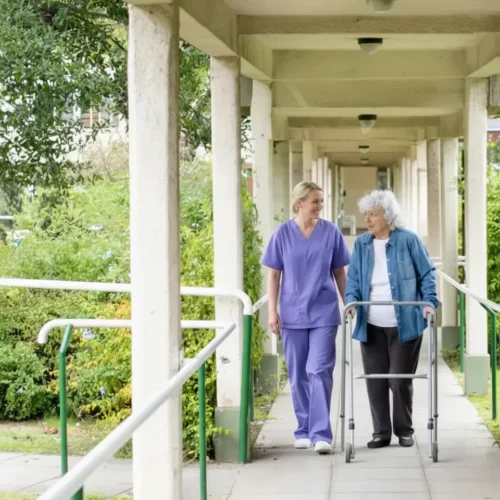 Nurse in purple scrubs walking outdoors with an elderly woman using a walker at an assisted living facility in Scottsdale.