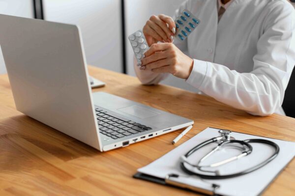 Close-up of a doctor holding blister packs of pills during an online consultation with a laptop and stethoscope on the desk.