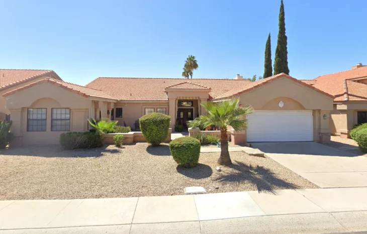 Front view of Assisted Living at Bloomfield Manor, Inc in Scottsdale, Arizona, showing a beige single-story home with a red tile roof, trimmed bushes, and palm trees under a clear blue sky.