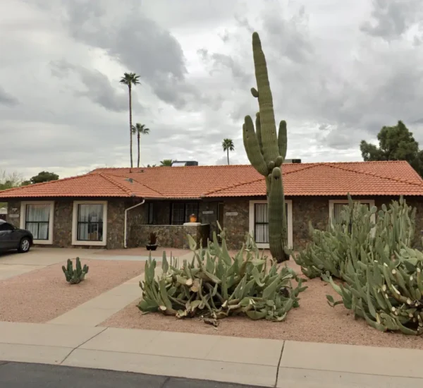 Single-story assisted living home in Scottsdale with a stone exterior, red tile roof, large cactus plants in the front yard, and a parked SUV in the driveway under cloudy skies.
