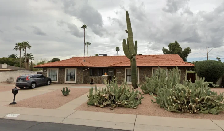 Single-story assisted living home in Scottsdale with a stone exterior, red tile roof, large cactus plants in the front yard, and a parked SUV in the driveway under cloudy skies.