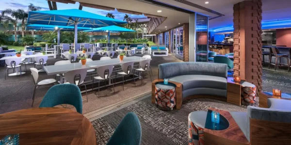 Open-air lounge at Hotel Valley Ho in Scottsdale with indoor seating, a bar area, and a patio dining space under bright blue umbrellas.