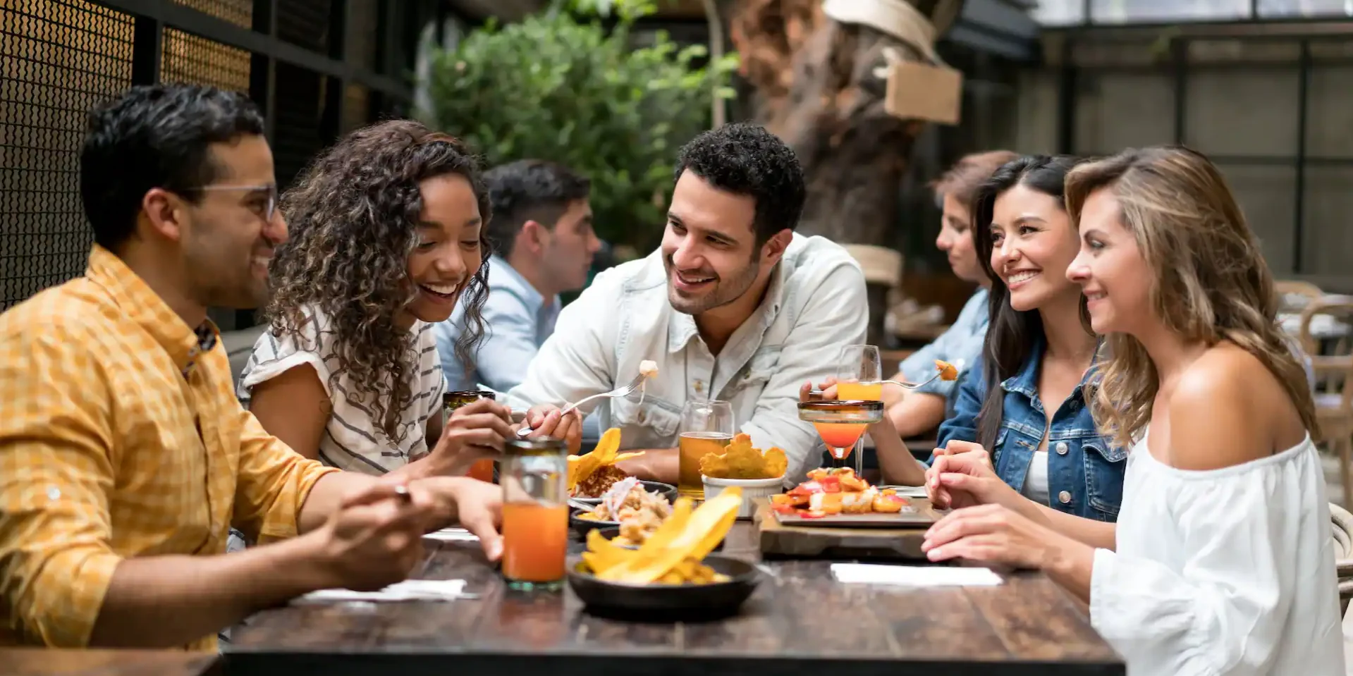 A warm, naturally lit lifestyle composition featuring a diverse group of friends gathered around a rustic dark wood table. The lighting is soft and golden, highlighting the condensation on glass jars filled with vibrant orange juice and the textures of a denim jacket and white off-the-shoulder top. The focus is on the joyful, candid expressions of the group as they share small plates of tropical food, including fried plantains. The background shows a softly blurred modern restaurant interior with industrial mesh accents and greenery.