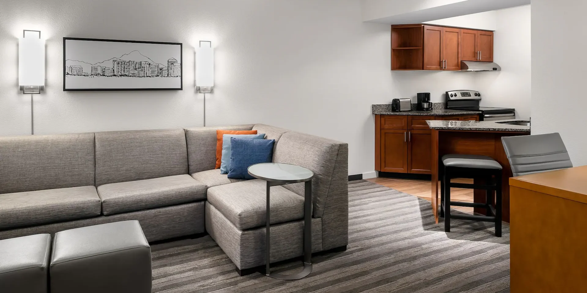 A clean, modern hotel suite interior showcasing a split composition between a living area and a kitchenette. The space is illuminated by warm, geometric wall sconces and recessed ceiling lights. On the left, a textured grey L-shaped sectional sofa is accented with bright orange and blue pillows. To the right, a compact kitchen features dark cherry-toned cabinetry, granite countertops, and sleek stainless steel appliances. The perspective is grounded by a grey and charcoal striped carpet that leads the eye toward the brightly lit kitchen corner.