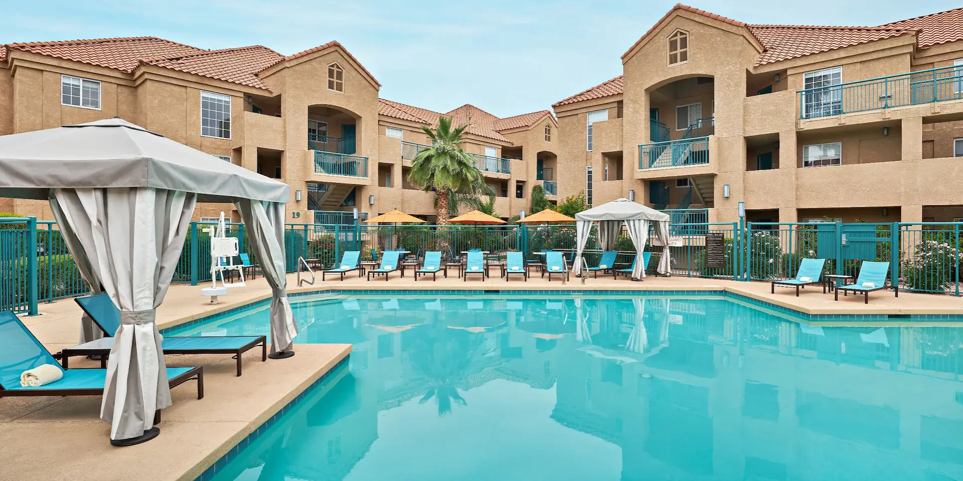 A wide, symmetrical architectural shot of a turquoise outdoor swimming pool centered within a multi-story, sand-colored stucco resort complex. The scene is bathed in bright, clear daylight under a soft blue sky. The composition features balanced rows of vibrant blue lounge chairs and cream-colored private cabanas with striped curtains. The pool's surface is exceptionally still, perfectly reflecting the building and a central palm tree. Crisp shadows on the pool deck provide a sense of depth and clean geometry.