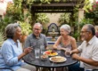 Bright, intense natural sunlight fills a lush Spanish-style courtyard (Mission/Pueblo architecture) featuring stone walls, potted desert plants (Agave, Saguaro), and a vibrant blooming bougainvillea. A horizontal composition captures four diverse senior adults laughing together around a centered, black wrought-iron mesh table. Sunlight casts distinct shadows on the patio floor. The table is laden with bowls of nuts, glasses of water, vibrant plates of fresh sliced watermelon and citrus fruit, and circular crackers on white plates. A decorative tiled fountain is visible in the background, all captured in warm, high-contrast natural daylight.