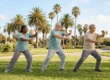 Wide horizontal shot of three diverse seniors in neutral-toned casual wear performing synchronized Tai Chi movements on a lush green lawn. The scene is bathed in bright, natural daylight with a soft-focus background of tall palm trees and a classic stone fountain under a clear blue sky in Scottsdale.