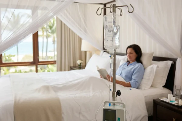 A sharp, detailed focus on a clear IV fluid bag and a digital infusion pump with a glowing green screen. The background is a soft-focus view of a luxury resort room with a canopy bed where a woman is comfortably reading a book. Bright, airy light suggests a tropical resort environment. The vertical lines of the IV stand create a strong focal point in the foreground.