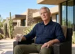 A cinematic photograph under harsh, natural daylight. Direct sunlight from the right highlights textures on an elderly man in a blue linen shirt and a patterned stone-and-glass building. He is seated centrally on a desert patio, holding a textured crystal glass. To the left, a background of Saguaro cacti and diverse desert flora against a vibrant blue sky is balanced by the modern architecture.