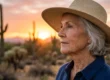The image is a medium close-up lifestyle photograph of a senior woman in an Arizona desert landscape during the golden hour sunset. She is shown in a right-side profile, looking towards the left, wearing a navy blue button-up shirt and a wide-brimmed woven straw hat which serves as sun protection. The lighting is warm and backlighting, coming from the low sun in the background, casting a soft glow on her silver hair and weathered skin, highlighting the texture of the straw weave. The background features blurred (bokeh) Saguaro cacti and mountains under a warm gradient sky. No text or branding is visible.
