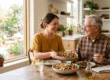 A warm, sunlit medium-shot composition bathed in soft, natural sunlight streaming from a large left-side window. The directional lighting highlights the vibrant, true-to-life colors of a fresh meal on a wooden table and creates a bright, uplifting tone. The shot utilizes a shallow depth of field, softly blurring the airy kitchen background to draw clear focus to the two smiling subjects in the center. There is no text visible in the frame.