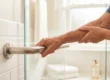A focused, horizontal photograph capturing the textured, wrinkled skin of an older person's forearm and hands firmly gripping a polished stainless steel safety grab bar. The person, wearing a blue shirt, has their hands positioned in the center, supported by each other. The metallic bar is mounted on a wall of clean, glossy white subway tiles on the left, with the surrounding bathroom softly blurred. Natural, warm light streams from a background window, highlighting the metallic sheen and the textures of the skin. Stacked white towels and a window are visible in the background bokeh.
